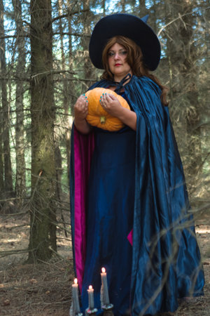 A red-haired witch in a pointed hat and a blue cloak in the forest with a large pumpkin in her hands burning candles on the ground. vertical photoの写真素材