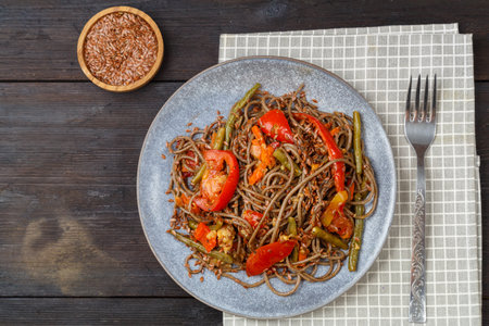 Soba with vegetables in soy sauce sprinkled with flax seeds in a plate on a napkin on a wooden table next to flax seeds in a saucer. horizontal photo.の写真素材