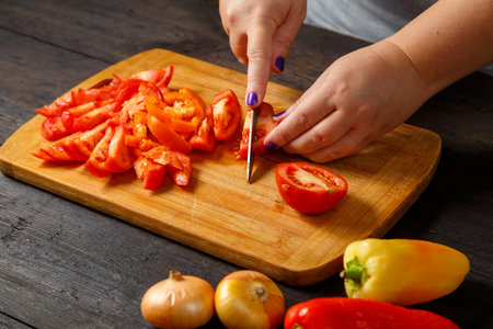 A woman in the kitchen with a knife cuts red tomatoes on a wooden board on the table next to onions and peppers. horizontal photoの写真素材