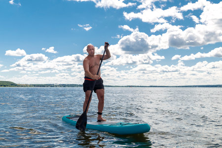 A man in shorts and a T-shirt on a SUP board swims in the lake on a sunny day against a sky with clouds. horizontal photoの写真素材