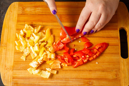 A knife in female hands cuts red sweet peppers on a wooden board into pieces. horizontal photoの写真素材