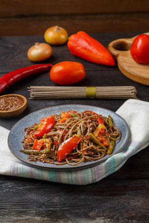 Soba with vegetables in soy sauce sprinkled with flax seeds in a plate on a napkin next to onions, tomatoes, peppers and flax seeds and uncooked soba on a wooden table. vertical photoの写真素材