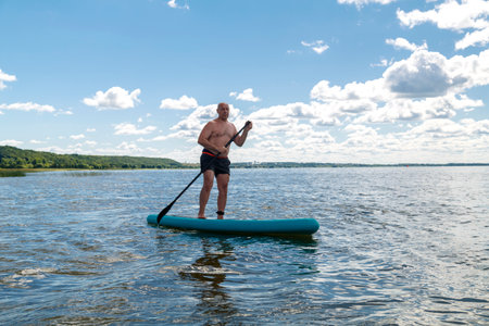 A man in shorts and a T-shirt on a SUP board in a lake on a sunny day against a sky with clouds. horizontal photoの写真素材