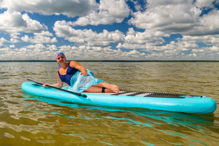 A woman with a Mohawk in a pareo lies sunbathing on a SUP board in a lake against the background of white clouds against a clear blue sky. horizontal photoの写真素材