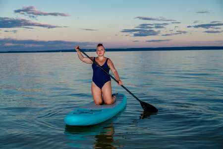 a woman in a closed swimsuit on her knees on a SUP board with an oar floats on the water against the background of the sunset sky. horizontal photoの写真素材