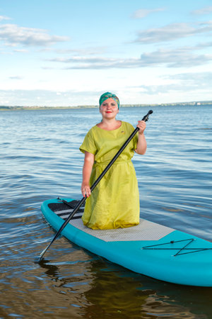 A Jewish woman in a green dress and a headscarf on her knees on a sup board against a clear blue sky. vertical photoの写真素材