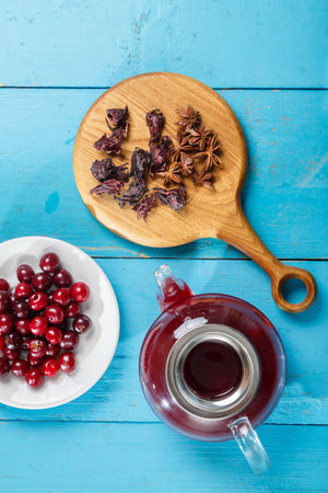 Kettle of hibiscus tea nearby on a blue table on a wooden board anise and cinnamon and hibiscus flowers and cherries in a plate top view. vertical photoの写真素材