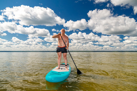A man in sports shorts with a paddle stands on a SUP board in the lake against the background of white clouds on a clear blue sky. horizontal photoの写真素材
