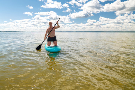 A man balances on a SUP board with a paddle in the lake on a sunny day against the backdrop of white clouds. horizontal photoの写真素材