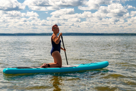 A woman in a swimsuit with a short haircut on a SUP board with a paddle in the lake against the background of white clouds. horizontal photoの写真素材