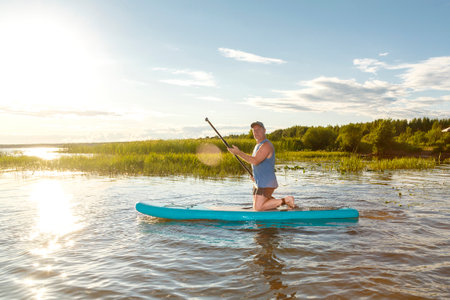 A man kneeling in a blue T-shirt on a SUP board with an oar in a lake near the reeds on a sunny day against the backdrop of white clouds. horizontal photoの写真素材