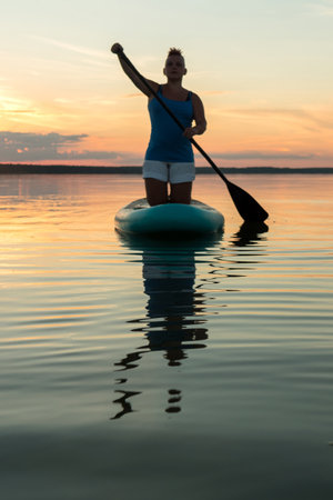 A woman with a mohawk in shorts on her knees on a sapboard with an oar against the backdrop of a sunset sky swims in the lake in the evening. vertical photoの写真素材