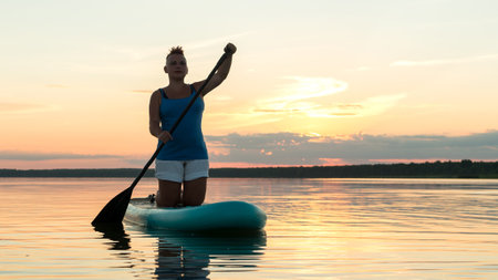 A woman on a sapboard on her knees with an oar at sunset against the sky floats in the water of the lake. horizontal photoの写真素材