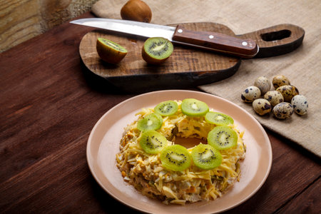 Layered salad with chicken and kiwi next to the board kiwi and quail eggs and a knife on the table. horizontal photoの写真素材