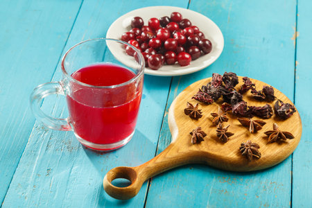A transparent cup of hibiscus tea next to the table on a wooden board anise and cinnamon and hibiscus flowers and cherries in a plate. horizontal photoの写真素材