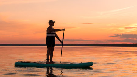 A man on a SUP board with a paddle at sunset against a pink-orange sky swims in the water of the lake. horizontal photoの写真素材