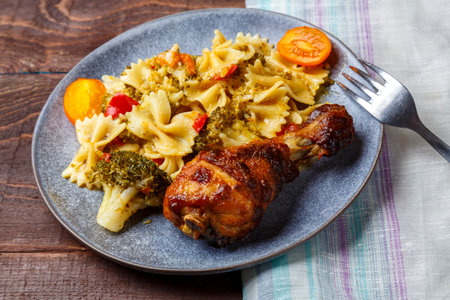 Pasta with vegetables and cherry tomatoes next to a baked chicken leg on the table next to a light napkin and a fork. horizontal photoの写真素材