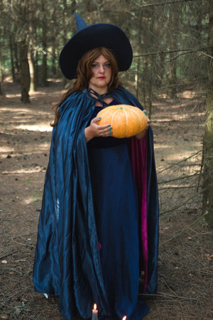 A red-haired witch in a pointed hat and a blue long cloak in the forest with a large pumpkin in her hands burning candles on the ground. vertical photoの写真素材