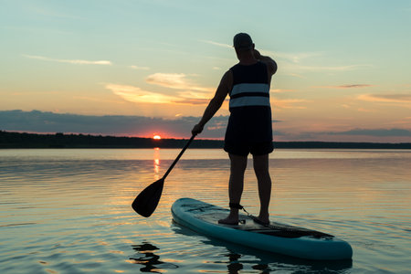 A man in shorts on a sapboard with an oar against the backdrop of a sunset sky swims in the lake in the evening. horizontal photo.の写真素材