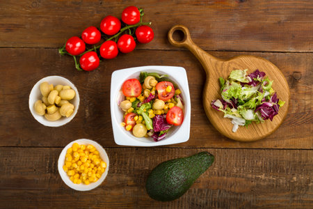 Vegetable salad with champignon corn and cherry tomato with avocado in a white plate on a wooden table next to all the ingredients. horizontal photoの写真素材