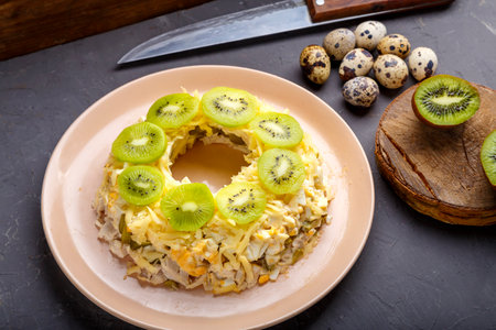 Layered salad with chicken and kiwi next to quail eggs kiwi and a knife on a gray table. horizontal photoの写真素材