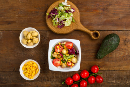 Vegetable salad with champignon corn and cherry tomato with avocado in a white plate on a wooden table next to the ingredients. horizontal photoの写真素材