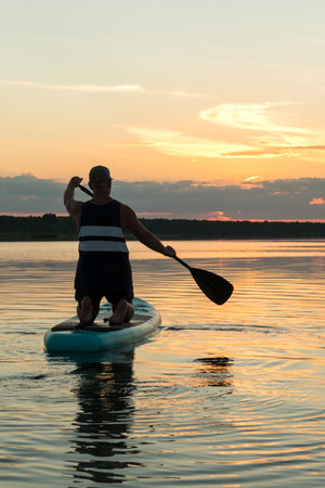 A man in shorts on a SUP board with an oar against the backdrop of a bright sunset sky swims in the lake in the evening. vertical photoの写真素材