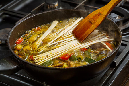 A wooden spatula mixes udon with stewed vegetables in spices in a frying pan. horizontal photo.の写真素材