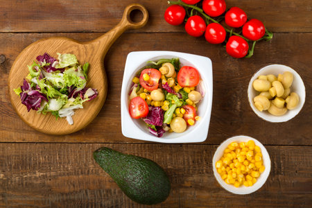 Vegetable salad with champignon corn and cherry tomato with avocado in a white plate on a wooden table next to all the ingredients. horizontal photo.の写真素材