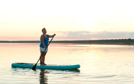 A woman in shorts and a top on a SUP board with a paddle at sunset swims in the water of the lake in the glare of the setting sun. horizontal photo.の写真素材