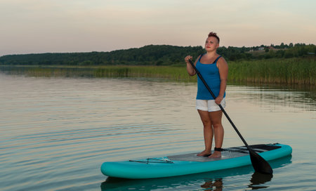 A woman in mohawk shorts stands on a SUP board at sunset in a lake against a pink and blue sky. horizontal photoの写真素材