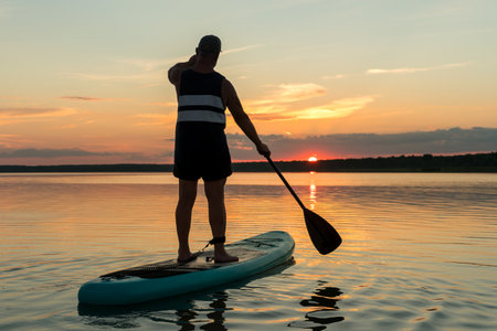 A man in shorts and a T-shirt on a SUP board with a paddle against the backdrop of the sunset sky in the lake. horizontal photoの写真素材