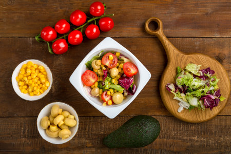 Vegetable salad with champignon corn and cherry tomato with avocado in a white plate on a wooden table next to all the ingredients. horizontal photoの写真素材