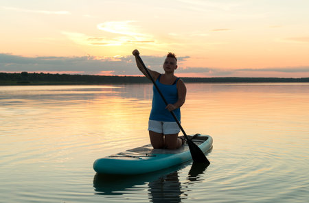 A woman on a sapboard on her knees with an oar at sunset against the sky floats in the water of the lake. horizontal photoの写真素材