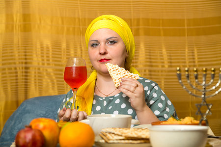 At the festive table on Shabbat, a Jewish woman in a Kisui Rosh headdress eats matzah after...