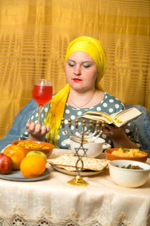 At a festive table with traditional food on Shabbat, a Jewish woman in a headdress with a glass of wine and a siddur makes kiddush. vertical photoの写真素材