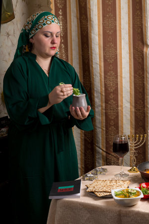 A woman in a green dress and headscarf at the table set for the Passover seder holds bitter herbs and a jug of salt water in her hands. vertical photoの写真素材