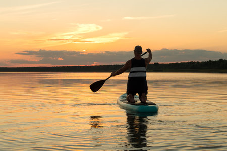 A man in shorts with his back to the camera on his knees on a SUP board with an oar swims in the lake against the background of the sunset sky. horizontal photoの写真素材