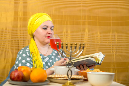 At the festive table with traditional food on Shabbat, a Jewish woman in a headdress with a glass of...