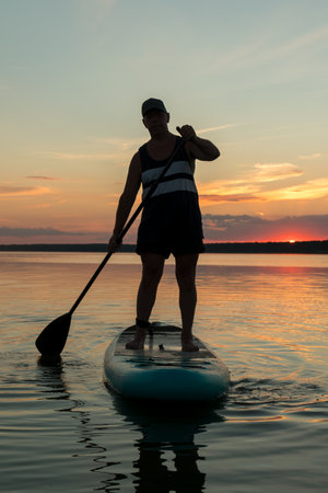 A man in shorts stands on a SUP board with a paddle in the evening at sunset in the lake. vertical planの写真素材