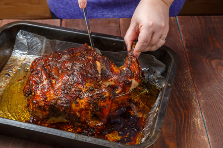 Women's hands tear off a leg from a whole baked chicken in a baking sheet with a knife. horizontal photo.の写真素材