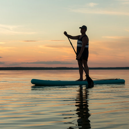 A man on a SUP board with a paddle swims in the calm water of the lake against the backdrop of the evening sky at sunset. square photoの写真素材