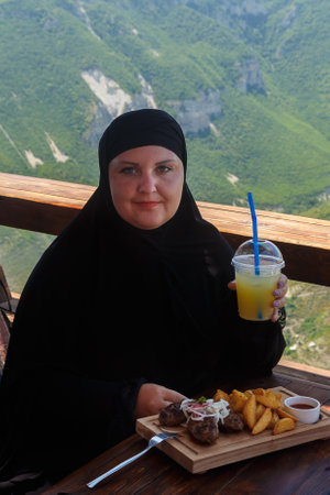 A Muslim woman in a black niqab sits on the veranda of a restaurant against the backdrop of mountains and drinks a cocktail. Vertical photoの写真素材