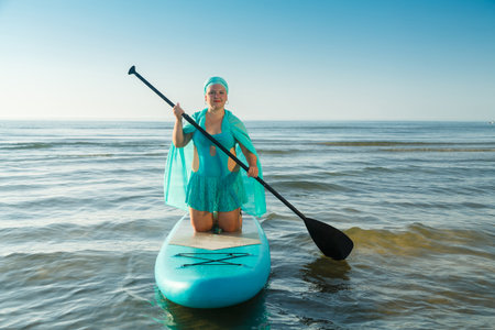 A woman in a turquoise swimsuit with a skirt and a scarf on her head on her knees on a SUP board swims to the seashore. horizontal photoの写真素材
