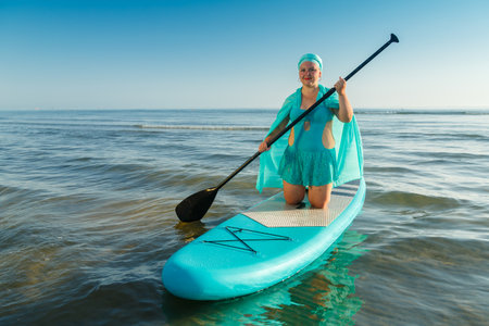 A Jewish woman in a turquoise bathing suit with a skirt and a headscarf on her knees on a SUP board sails away from the seashoreの写真素材