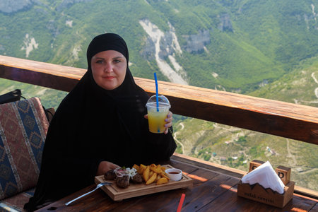 A Muslim woman in a black niqab sits on the veranda of a restaurant against the backdrop of mountains and drinks a cocktail. Horizontal photo.の写真素材
