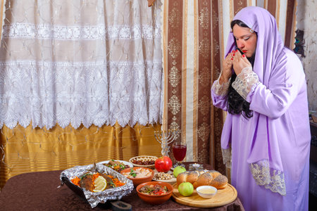 A Jewish woman in a wig and a veil falling from her hair and a long dress reads a blessing on the candles on Rosh Hashanah, Shabbat at the set table. Horizontal photo.の写真素材