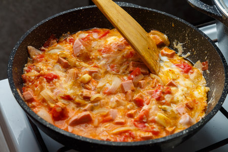 Shakshuka with tomatoes and sweet cheese sprinkled with a wooden spatula in a frying pan on the stove. horizontal photoの写真素材