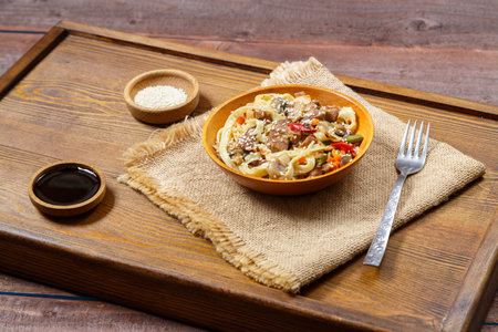 Wok udon noodles with beef and vegetables in a wooden plate on a salad on a hearth board next to soy sauce and sesame seeds. Horizontal photo.の写真素材