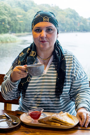 A Jewish woman in a headscarf drinks tea with pancakes against the backdrop of a window overlooking the lake. Vertical photoの写真素材